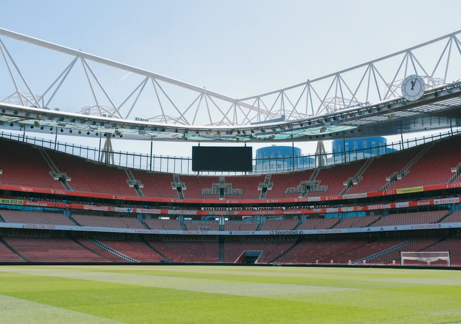 An empty sports stadium with a well-maintained green grass pitch, surrounded by red seating sections and modern white structural beams supporting a partially covered roof. The stadium features digital screens and a large clock visible at the upper level, with bright natural daylight illuminating the space. The seating area appears clean and organized, reflecting a maintained environment suitable for hosting sporting events, as part of a professional cleaning and maintenance process by Islington Carpet Cleaning, aligning with surface cleaning and hygiene standards for commercial spaces.