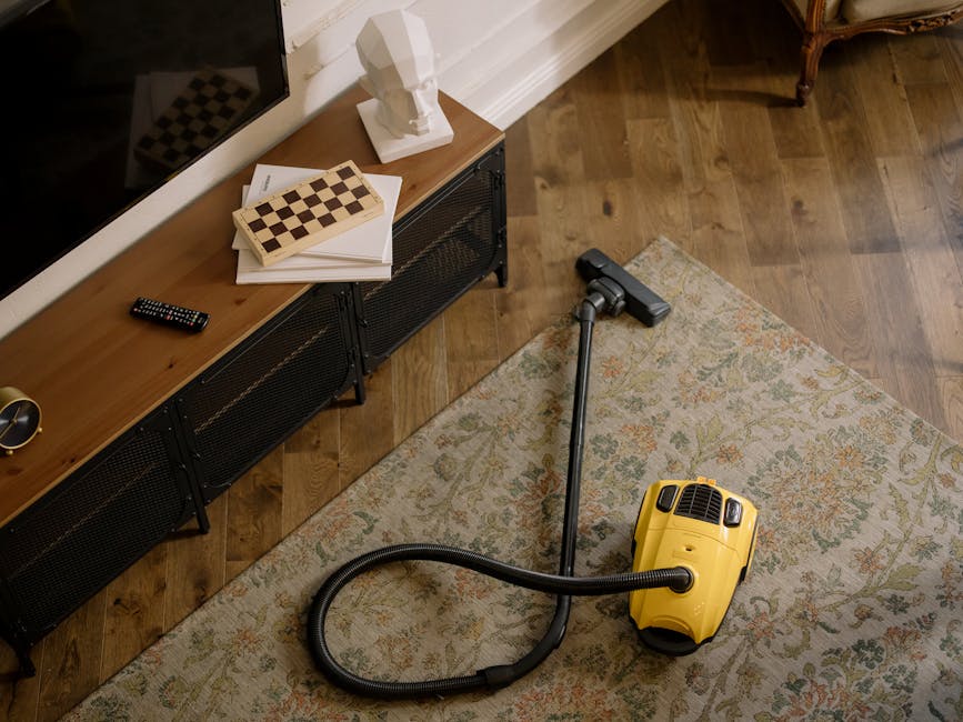 A close-up view of a living room corner featuring a hardwood floor with an ornate patterned area rug. On the floor, there is a yellow handheld vacuum cleaner connected to a black vacuum hose. Adjacent to the rug, a long black vacuum wand extends from the cleaner. In the background, a wooden sideboard with a black metal frame holds a white bust sculpture, a remote control, a stack of papers, and a chessboard. The room is well-lit, emphasizing the clean and polished surfaces of the wooden floor and the tidy appearance of the space, illustrating typical surface cleaning and maintenance with a focus on domestic cleaning at Islington Carpet Cleaning.