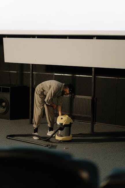 A person dressed in a beige jumpsuit is using a yellow wet vacuum cleaner on a dark carpeted floor in a room with black wall panels and a white suspended ceiling. The vacuum hose is extended across the floor, indicating a process of deep cleaning or surface sanitisation. Visible equipment includes a large speaker placed against the wall and a window with a radiator underneath. The room appears clean and organized, with a professional setting suitable for commercial or residential carpet cleaning services, as offered by Islington Carpet Cleaning. The lighting is even, highlighting the cleaning procedure in progress.