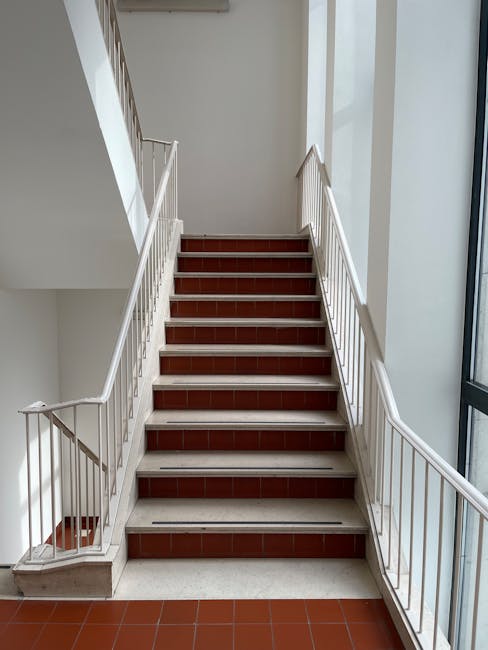 Photograph of a narrow staircase inside a building, featuring white-painted metal railings on both sides and red tile steps. The stairway is well-lit by natural light coming from large windows on the right, revealing a clean, dust-free surface. The surrounding walls are painted white, contributing to the bright and tidy appearance. The flooring at the base is covered with matching red tiles, and the overall scene suggests a well-maintained interior space. The image is associated with Islington Carpet Cleaning and relates to difficult access cleaning in narrow stairs, emphasizing the importance of thorough surface cleaning and sanitisation of confined staircase environments in residential or commercial settings.