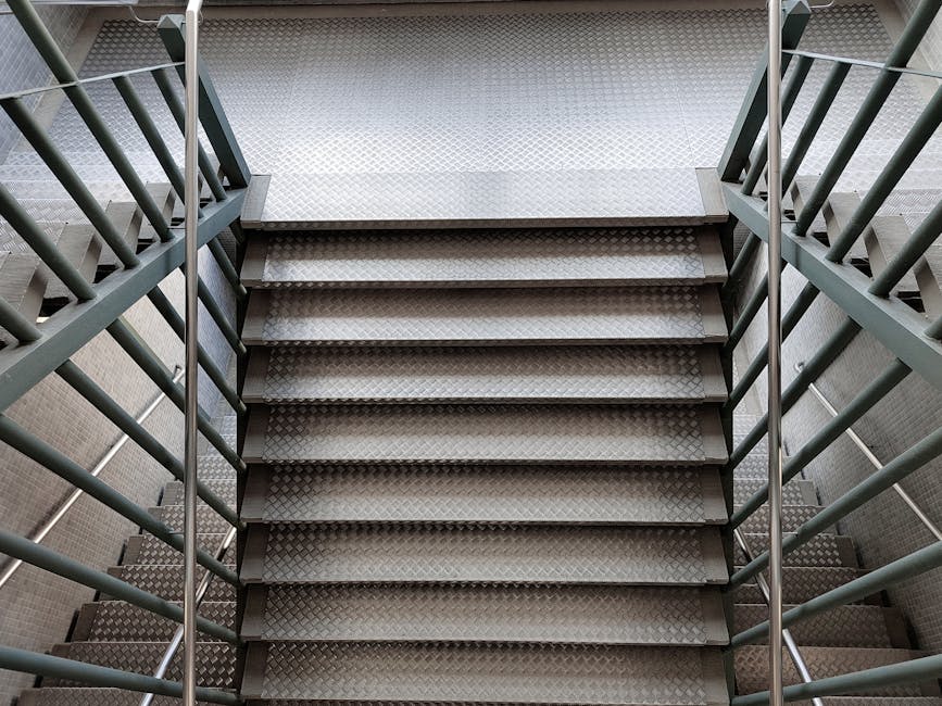 Close-up view of a narrow metal staircase with diamond-patterned anti-slip treads, flanked by green-painted metal railings on both sides. The steps appear slightly worn but clean, with no visible dust or debris, in a well-lit indoor setting. The surrounding walls are tiled in a neutral color, and natural light illuminates the scene from above, highlighting the surface texture and metallic shine. This image exemplifies surface cleaning and maintenance of challenging access areas, reflecting the expertise of Islington Carpet Cleaning in deep cleaning hard-to-reach spaces like narrow stairs in residential or commercial buildings.