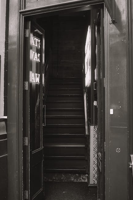 Photograph of a narrow staircase inside a building, featuring white-painted metal railings on both sides and red tile steps. The stairway is well-lit by natural light coming from large windows on the right, revealing a clean, dust-free surface. The surrounding walls are painted white, contributing to the bright and tidy appearance. The flooring at the base is covered with matching red tiles, and the overall scene suggests a well-maintained interior space. The image is associated with Islington Carpet Cleaning and relates to difficult access cleaning in narrow stairs, emphasizing the importance of thorough surface cleaning and sanitisation of confined staircase environments in residential or commercial settings.