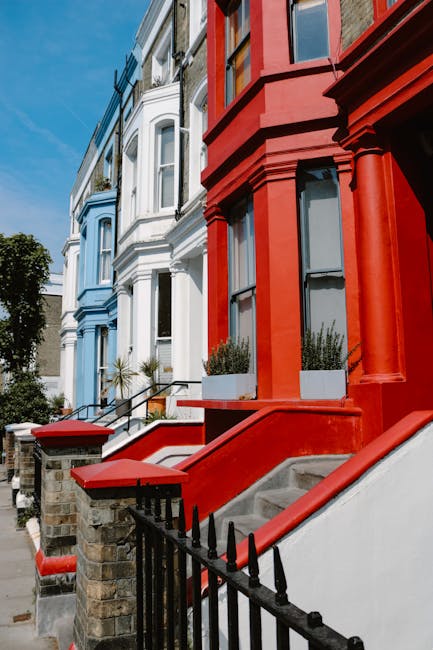 Image of a row of colorful Victorian-style residential buildings along a sunny street in Islington, London. The buildings feature detailed facades with bay windows, decorative moldings, and vibrant colors—one in bright red and others in pastel blue and white. Each building has steps leading up to its entrance, with black wrought iron railings and small front gardens with potted plants. The street is lined with a clear blue sky and some trees in the background. The scene emphasizes well-maintained exteriors and the urban architectural charm typical of Islington. This image is associated with Islington Carpet Cleaning's domestic cleaning services, including surface cleaning and deep cleaning to maintain property hygiene.