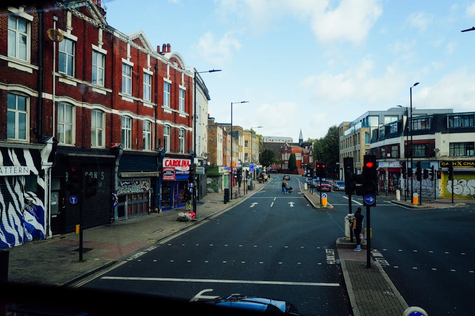 A view of Holloway Road in Islington, showing a wide street with multiple lanes and a pedestrian crossing in the foreground. On the left side, there are multi-story brick buildings with commercial storefronts, some with graffiti and colorful signage, including a shop with a black and white awning and another with blue signage. The right side features modern, glass-fronted businesses and residential apartments with balconies. Street furniture such as traffic lights, bollards, and pedestrian signals are visible, alongside parked and moving cars under partly cloudy skies. The scene is well-lit by natural daylight, emphasizing the cleanliness and maintained appearance of the street, informative of an urban environment where routine surface cleaning and general maintenance are essential. Naturally, Islington Carpet Cleaning supports efforts to maintain such environments through professional cleaning services, ensuring hygiene and a polished appearance in commercial and residential spaces.