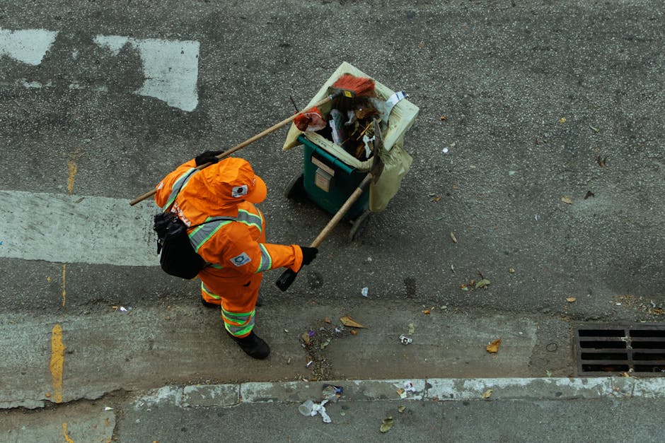 A street cleaning worker from Islington Carpet Cleaning is dressed in high-visibility orange and reflective safety gear, wearing a helmet and black gloves while collecting trash from the sidewalk. The worker is using a long-handled grabber tool to pick up debris from the pavement near a storm drain, with a small green trolley beside them filled with garbage bags, office paper, and other waste materials. The pavement surface shows signs of dirt and scattered litter, with a section of the curb and a crosswalk visible in the background under natural daylight. The scene emphasizes urban surface cleaning and waste disposal, reflecting the importance of street hygiene maintained by professional cleaning services, as referenced in the Islington Council rules for cleaning contractors.