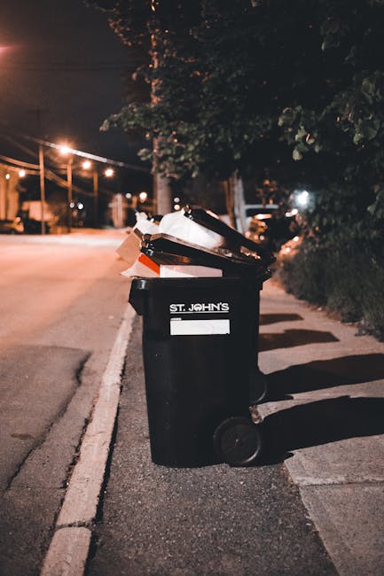 A row of several black outdoor waste bins with orange and blue lids, neatly aligned against a red brick wall. The bins are clean and closed, positioned on a paved surface, with natural daylight illuminating the scene. The setting appears to be an urban or residential area, emphasizing waste disposal and sanitation practices. This image reflects cleanliness and proper waste management, consistent with the services provided by Islington Carpet Cleaning as part of surface cleaning and hygiene maintenance in line with Islington Council rules for cleaning contractors waste disposal.