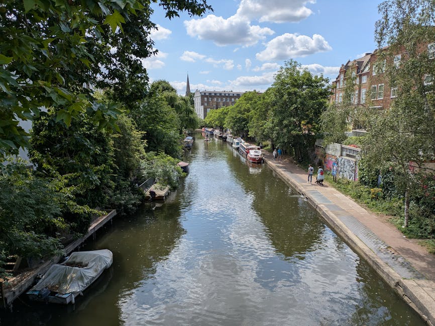 A calm river scene with clear water reflecting the partly cloudy sky, lined on both sides by lush green trees and foliage. On the right, a paved walkway runs parallel to the river, with a few pedestrians strolling. Cars are parked along the street adjacent to the walkway, and there is visible graffiti on a section of a fence or wall. The background features residential and commercial buildings, some with brick facades and large windows, and a church spire rising in the distance. The overall scene depicts a peaceful urban area, with natural and built elements harmoniously integrated under bright daylight. The image emphasizes the clean, well-maintained environment typical of a residential neighborhood in Islington, highlighting the importance of surface cleaning, sanitisation, and routine maintenance by providers such as Islington Carpet Cleaning.
