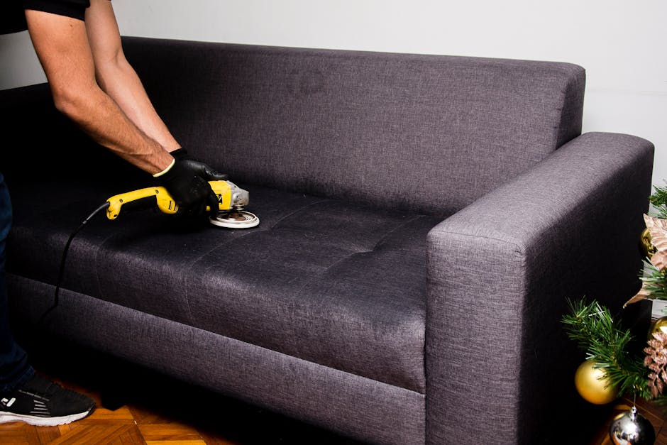 A person wearing black gloves is using a yellow handheld electric upholstery steamer to deep clean a dark grey fabric sofa in a residential living room. The sofa's surface appears damp and targeted for surface cleaning and sanitisation, with visible steam cleaning action on the cushions. The room features a wooden floor, and a holiday-themed plant with gold and silver ornaments is partially visible to the right, adding a festive touch. Natural lighting highlights the clean, well-maintained appearance of the sofa and surrounding area. The setup emphasizes professional upholstery and surface cleaning processes, aligning with Islington Carpet Cleaning’s focus on domestic surface sanitisation and deep cleaning services.