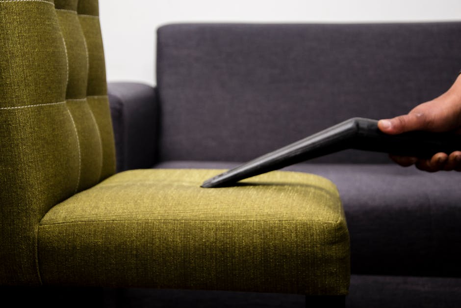 A close-up view of a person using a vacuum cleaner attachment to clean a green upholstered chair with tufted backrest cushions. The chair's fabric appears clean and well-maintained, situated in a room with a grey fabric sofa visible in the background. The surface of the chair shows no visible dust or dirt, indicating thorough surface cleaning and maintenance by Islington Carpet Cleaning. The lighting in the room is bright, highlighting the texture of the fabric. The use of professional cleaning tools suggests a focus on hygienic and deep cleaning processes, aligning with surface sanitisation practices for upholstery care.