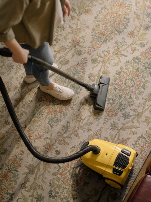 A person vacuuming an intricately patterned, multicolored carpet in a room with soft, natural lighting. The individual is wearing jeans, beige shoes, and a jacket, holding a hose attached to a yellow vacuum cleaner positioned on the floor. The carpet features shades of beige, green, and orange with floral motifs, and appears clean and well-maintained. Visible furniture includes a wooden table and a leather armchair. The scene illustrates surface cleaning and deep cleaning processes, reflecting hygiene and maintenance practices associated with domestic cleaning services provided by Islington Carpet Cleaning, as referenced on the N1 apartment carpet cleaning tips page for Angel locals on islingtoncarpetcleaning.org.uk.