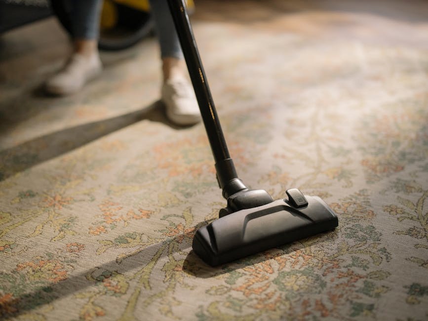 A person vacuuming an intricately patterned, multicolored carpet in a room with soft, natural lighting. The individual is wearing jeans, beige shoes, and a jacket, holding a hose attached to a yellow vacuum cleaner positioned on the floor. The carpet features shades of beige, green, and orange with floral motifs, and appears clean and well-maintained. Visible furniture includes a wooden table and a leather armchair. The scene illustrates surface cleaning and deep cleaning processes, reflecting hygiene and maintenance practices associated with domestic cleaning services provided by Islington Carpet Cleaning, as referenced on the N1 apartment carpet cleaning tips page for Angel locals on islingtoncarpetcleaning.org.uk.