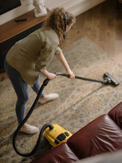 Close-up view of a vacuum cleaner with a black handle and head, being used to clean a patterned area rug in a well-lit living room; the rug features intricate floral designs in pastel shades, and the flooring underneath appears to be dark hardwood. The vacuum's attachment is in contact with the carpet's surface, indicating surface cleaning or deep cleaning process, with some evidence of dust or dirt removal. In the background, a person's legs and white sneakers are visible, suggesting a domestic setting. The scene emphasizes hygiene and maintenance, aligning with professional cleaning services offered by Islington Carpet Cleaning, as depicted on their website page discussing the cost of carpet cleaning in Islington N1.