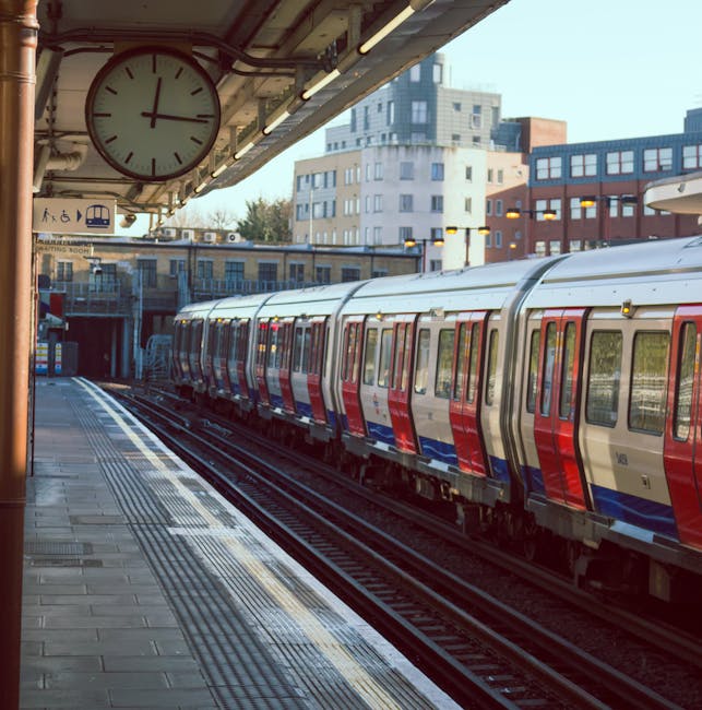 An outdoor railway station platform with a modern, clean train featuring a red, blue, and silver exterior stopped at the platform. The platform is paved with grey tiles, appearing tidy and well-maintained, with tactile paving along the edge for safety. Part of a large clock showing the time, mounted under the station's roof, is visible in the top left corner, along with some ceiling lighting. In the background, numerous multi-storey residential buildings with a mix of white and red facades are visible in the sunny daylight. The area looks orderly and bright, highlighting the importance of surface cleaning and maintenance in public transit environments, which [COMPANY_NAME] in Islington specializes in for ensuring hygiene and safety during busy hours.