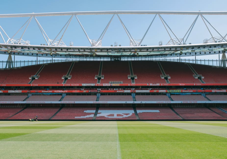 Inside a large football stadium with a well-maintained grass pitch in the foreground, showing the seating area with red seats and white lettering spelling out 'Arsenal' on the middle tier. The upper structure features a white, modern roof framework supported by steel trusses, with bright natural light illuminating the scene. The stadium appears clean and organized, reflecting high standards of surface cleaning and maintenance by Islington Carpet Cleaning, although specific tools or cleaning products are not visible in the image.
