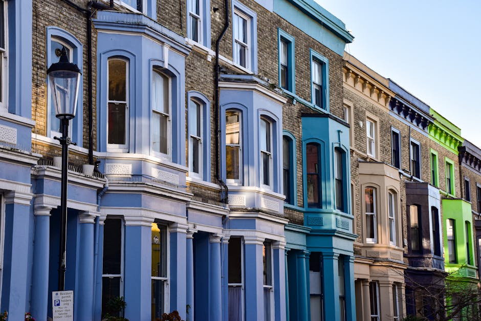 A row of Victorian-style terraced houses with colorful facades, including shades of light blue, beige, dark blue, and bright green, situated along a street illuminated by a vintage-style street lamp. The buildings have large, white-framed sash windows, some with decorative moldings, and feature brick and stucco exteriors. The clear sky above suggests daytime. This exterior scene highlights urban residential architecture typical of Islington, and reflects a well-maintained, clean appearance, consistent with professional cleaning services by Islington Carpet Cleaning for a neat and welcoming streetscape.