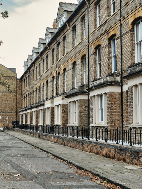 Photograph of a multi-storey residential building made of beige and brown brick, with a row of tall, narrow white-framed windows on each floor. The building features a horizontal decorative brick pattern beneath the windows and a slate roof with dormer windows. A black metal railing runs along the small front yard, which is paved with concrete slabs. The adjacent sidewalk is made of concrete, with a few fallen leaves scattered across it, and the scene is illuminated by natural daylight, suggesting an overcast sky. The overall appearance indicates a well-maintained, typical London townhouse exterior. This image is associated with residential cleaning and maintenance efforts by Islington Carpet Cleaning for ensuring clean, hygienic property exteriors in the Islington area.