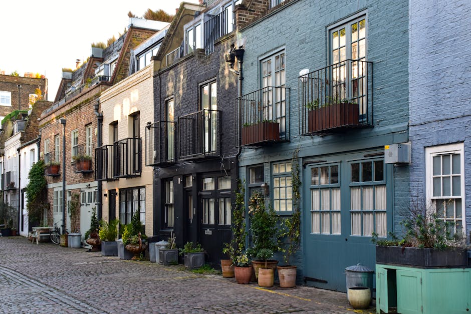 A row of Victorian-style terraced houses with colorful facades, including shades of light blue, beige, dark blue, and bright green, situated along a street illuminated by a vintage-style street lamp. The buildings have large, white-framed sash windows, some with decorative moldings, and feature brick and stucco exteriors. The clear sky above suggests daytime. This exterior scene highlights urban residential architecture typical of Islington, and reflects a well-maintained, clean appearance, consistent with professional cleaning services by Islington Carpet Cleaning for a neat and welcoming streetscape.